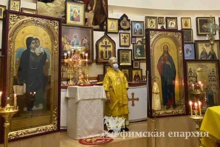 Митрополит Никон в жёлтом облачении перед престолом домового храма, рядом диакон в медицинской маске держит потир, вокруг — стены, увешанные иконами