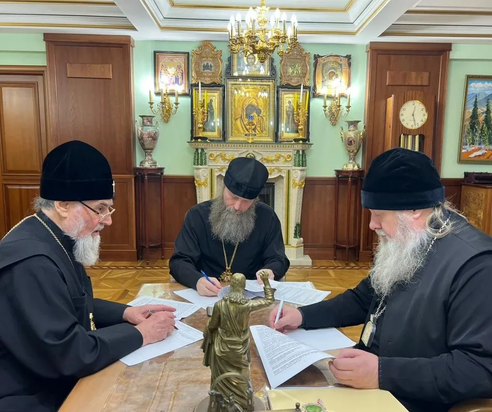 Три архиерея в чёрных облачениях подписывают документы за столом в кабинете, на заднем плане — угол комнаты с иконами в окладах
