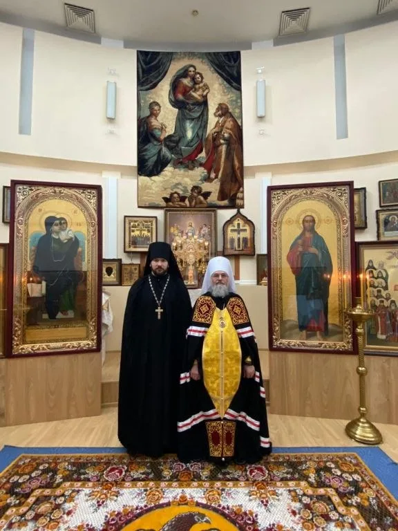 Митрополит Никон в чёрной мантии с жёлтой епитрахилью рядом с новопостриженным монахом на фоне иконостаса и образа Покрова Богородицы
