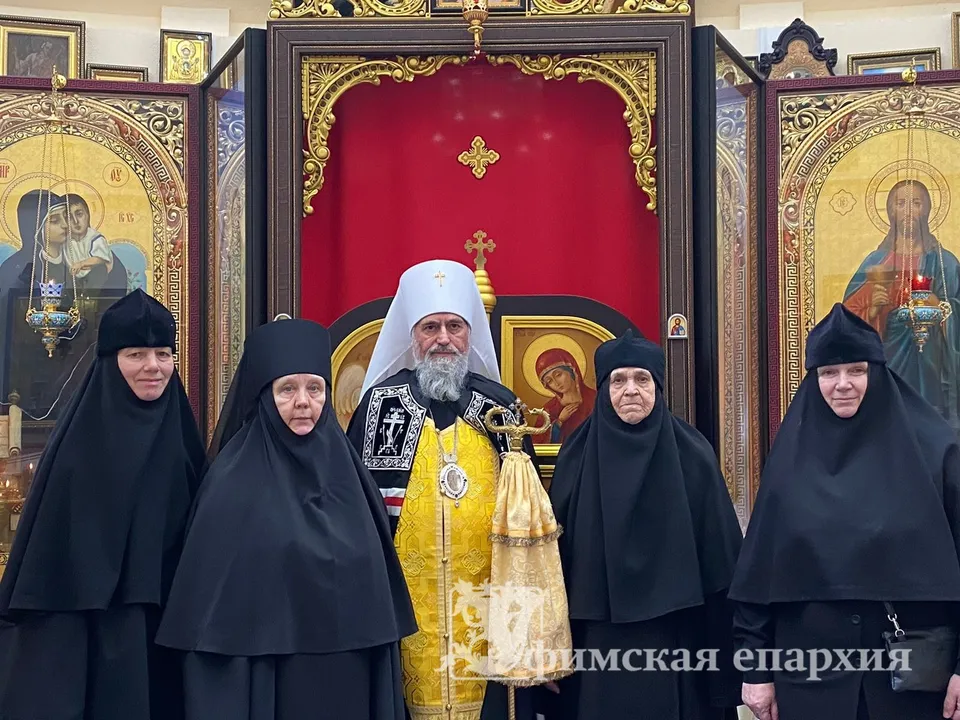 Митрополит Никон в жёлтом облачении с четырьмя монахинями в чёрных одеяниях перед резным золочёным иконостасом