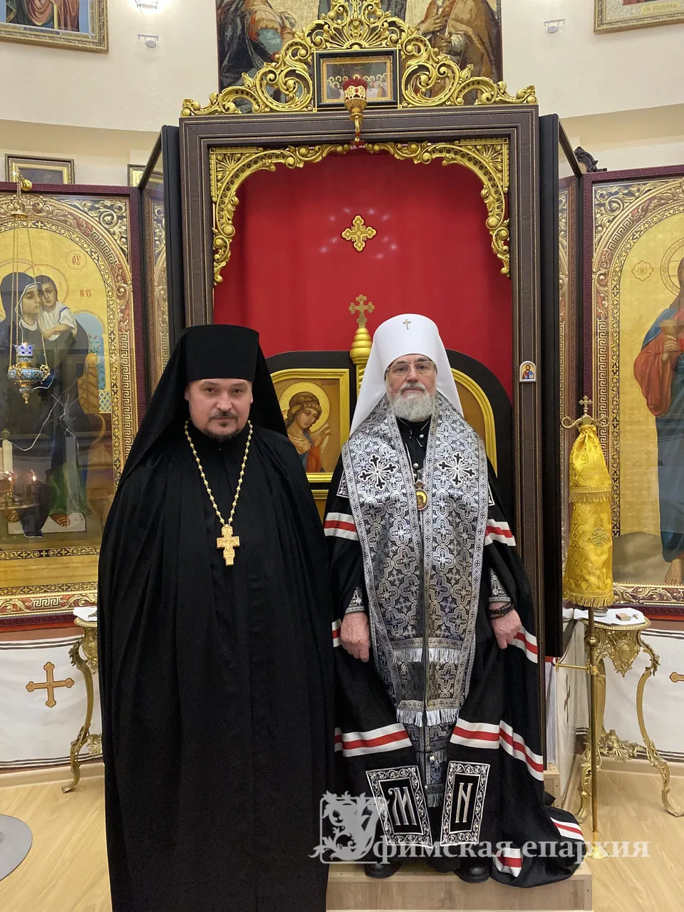 Митрополит Никон и новопостриженный монах стоят у раскрытого киота с образом Божией Матери на фоне рам с меднолитыми крестами и иконами