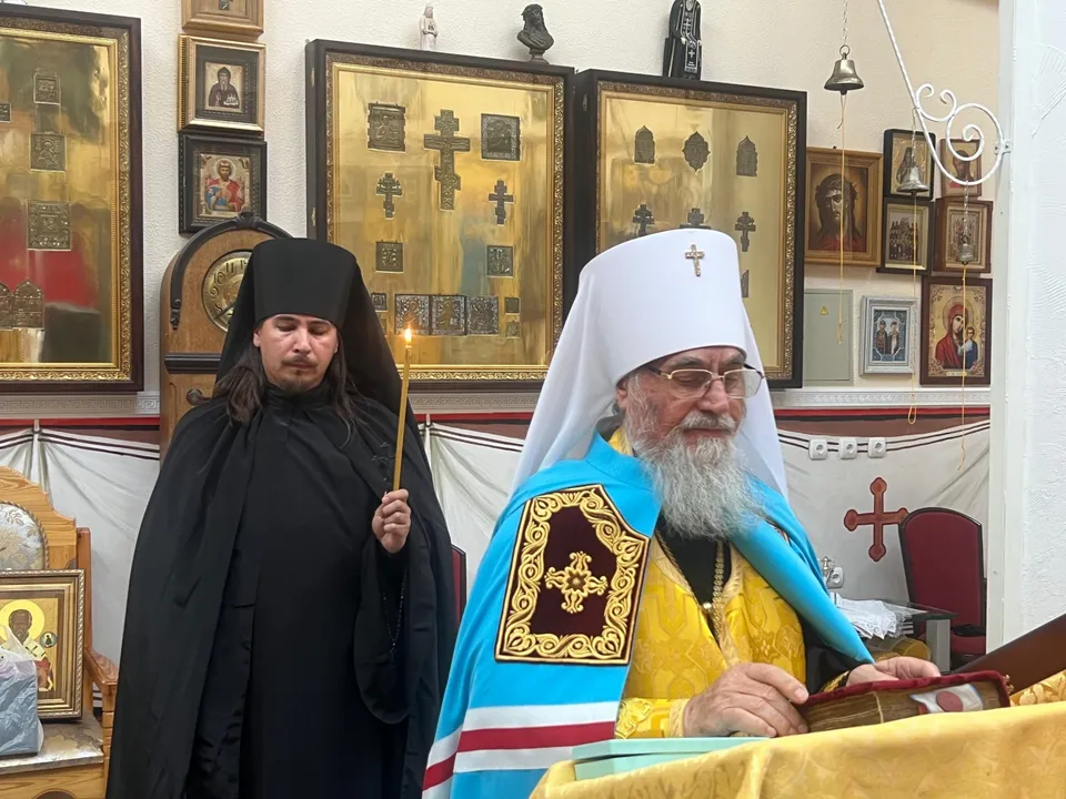 Митрополит Никон и молодой монах в чёрном облачении перед стеной с меднолитыми крестами и иконами разных размеров